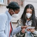 Focused young multiethnic female classmates in casual clothes and medical masks using tablet together while standing near brick building