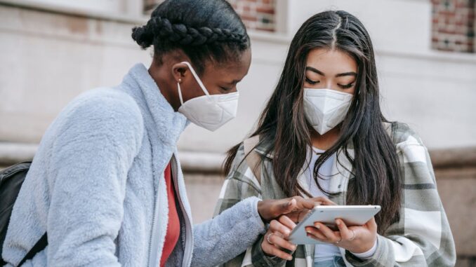 Focused young multiethnic female classmates in casual clothes and medical masks using tablet together while standing near brick building