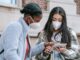 Focused young multiethnic female classmates in casual clothes and medical masks using tablet together while standing near brick building